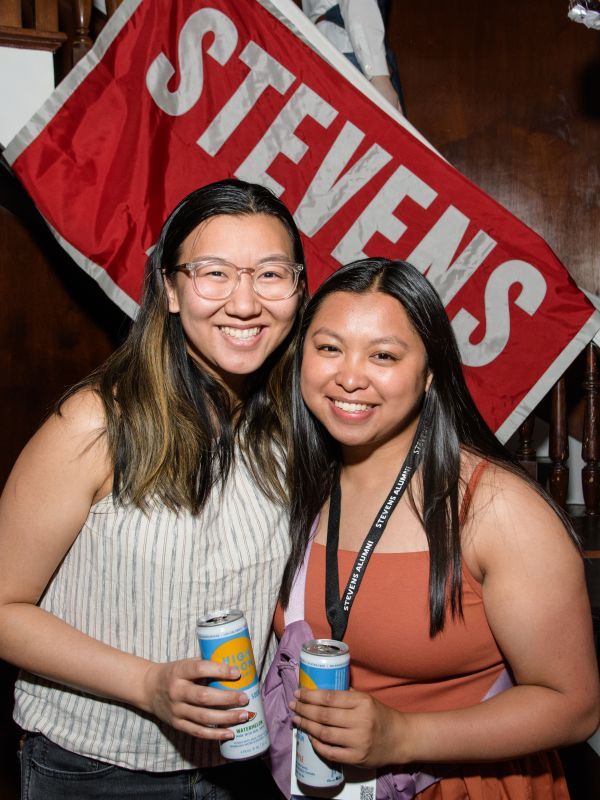 Two young alumni celebrate at Alumni Weekend in front of Stevens banner and balloons