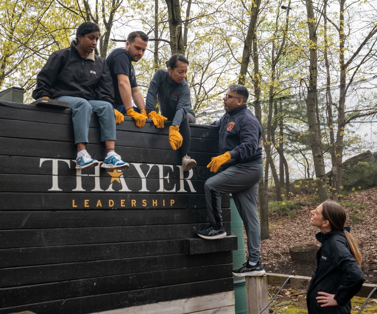 Four EMBA students sit on top of an obstacle course all with the Thayer Leadership logo discussing what their next move should be. 
