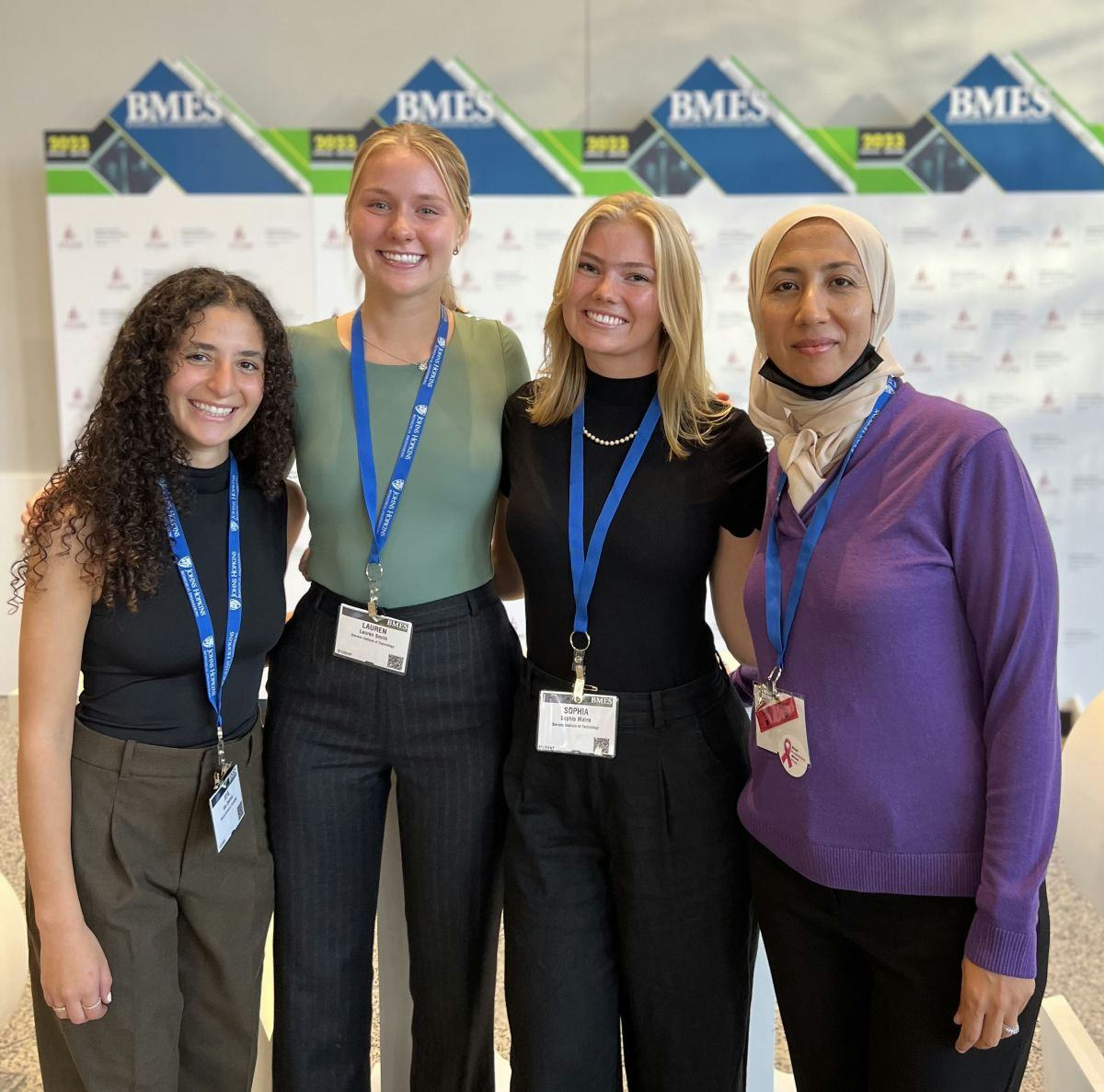 A group of four women stand together in front of a conference poster with "BMES" signage