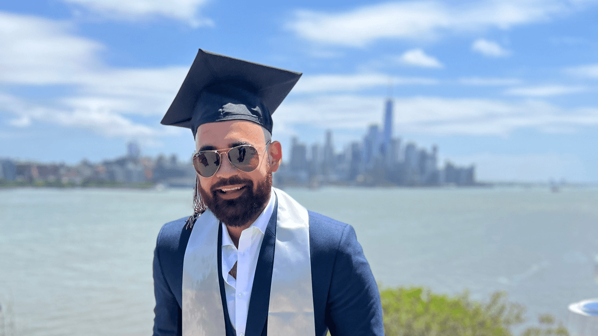 Rohan Chathrath stands with the New York City skyline in the background, wearing his graduation cap and sash.