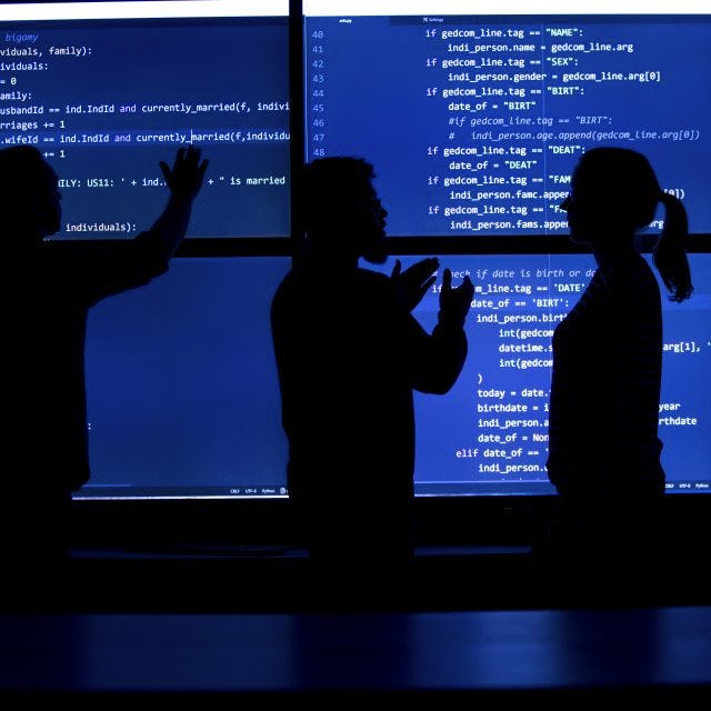 A silhouette of three people looking at a blue screen in a dark room.