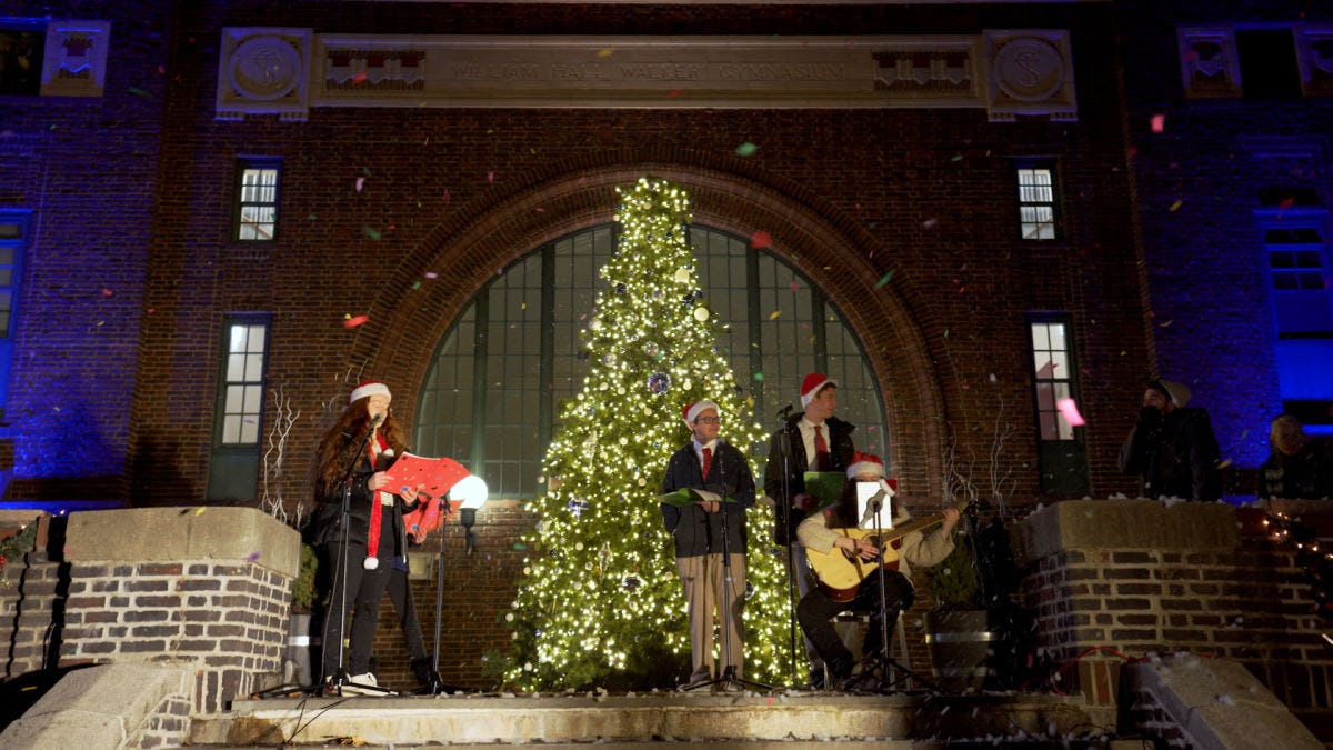 A band plays in front of a lit Christmas tree in front of Walker Gym.