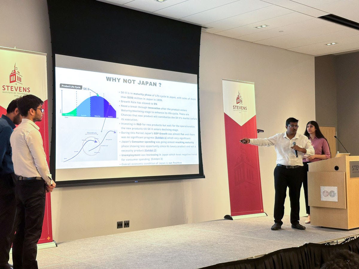 Tilak Chandana holds a microphone pointing at a PowerPoint slide while he stands on stage with his team at the Business Case Study Competition.