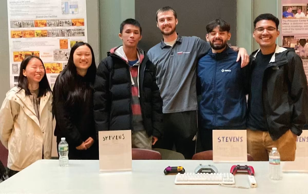 A group of six students stand behind a table wearing jackets. On the table is a keyboard and gaming remotes. Behind them are research posters.