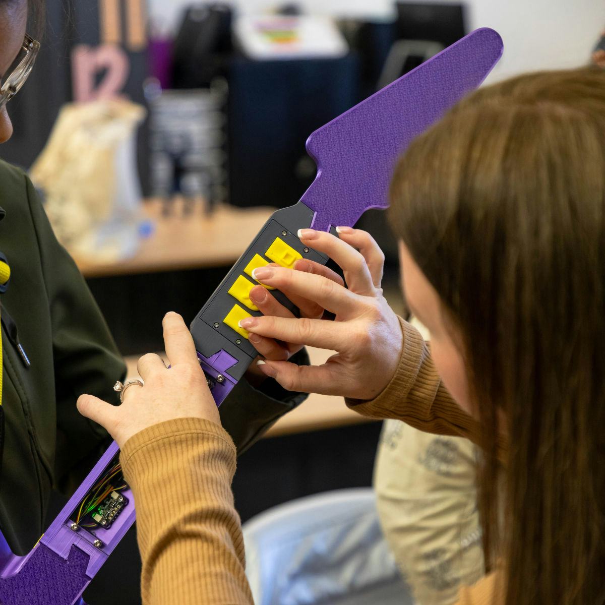 A student on the left plays a purple electric guitar and is guided by a teacher who helps her press the correct guitar buttons of  the Haptic Hero game. 