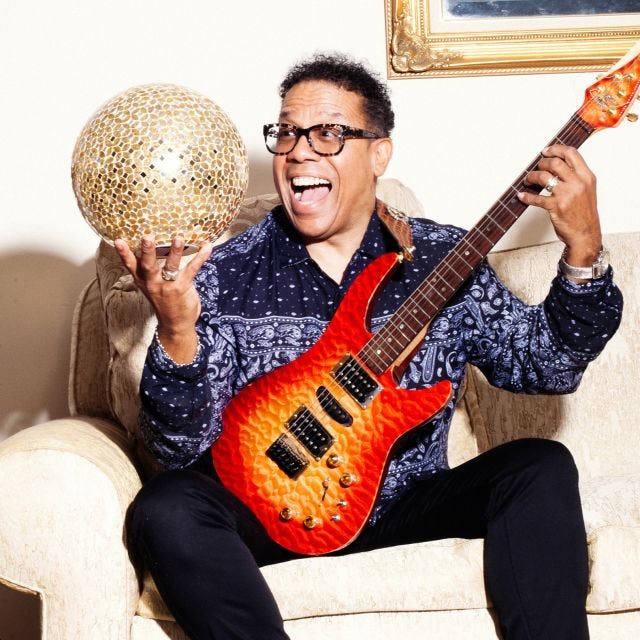 Carlos Alomar joyfully sits on a couch with his guitar and a golden orb.