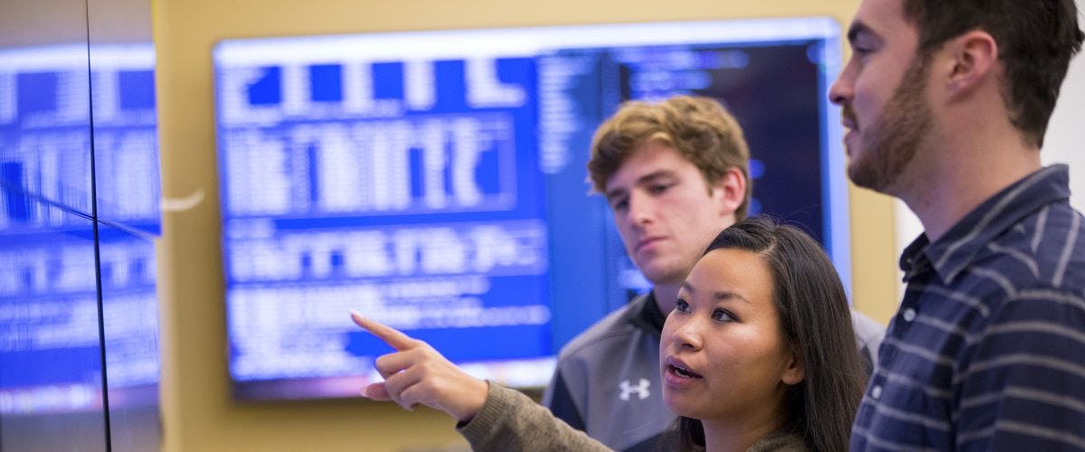 Student points at wall monitor while two other students look on.