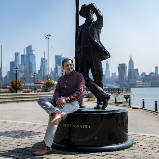 Mani sits on the Frank Sinatra statue in Hoboken with part of the New York City skyline behind him. 