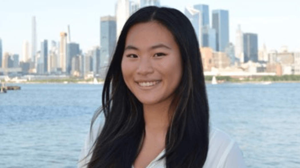 A photo of Katherine Ho standing before the Hudson River with the Manhattan skyline behind her