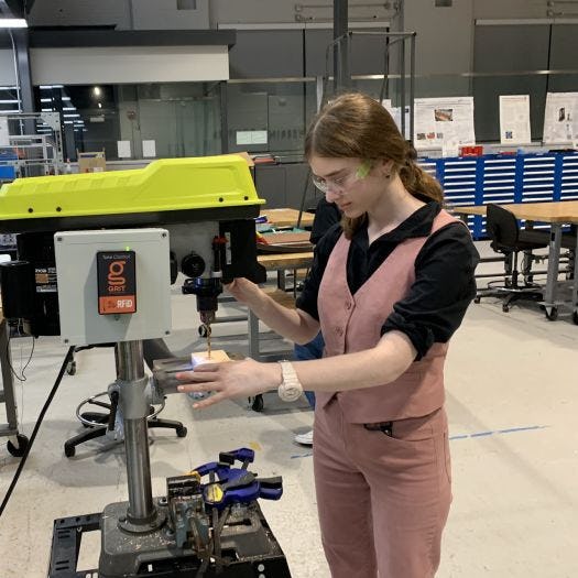 A female student wearing goggles stands in front of a machine she is operating, while holding an object in her hands that she is building.