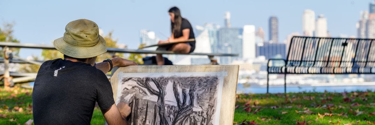 Student sits in grass and draws NYC skyline.